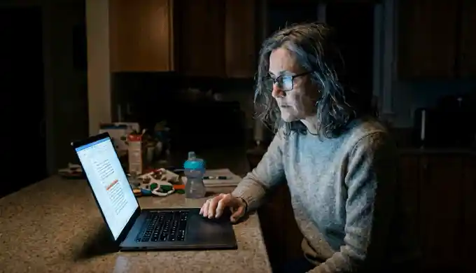 Dana reading research on a laptop at a kitchen table late at night.